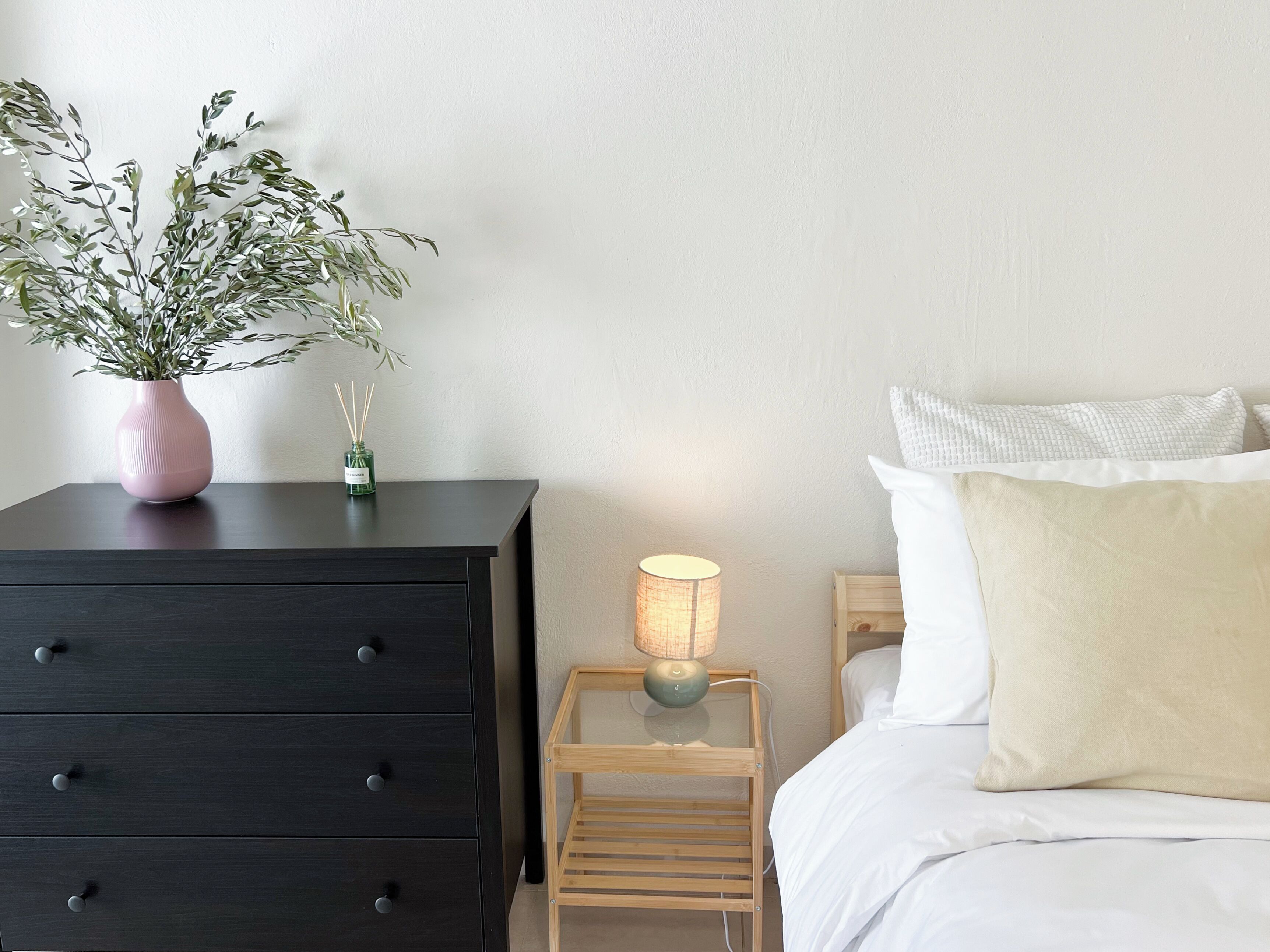 Een slaapkamer met een donker houten dressoir, een nachtkastje met een lamp, een klein houten krukje, en een bed met wit beddengoed en een beige kussen.