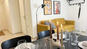 Ein Essbereich mit Tisch, gedeckt für vier Personen, inklusive Kerzen, Gläsern und Besteck. Im Hintergrund sind ein gelbes Sofa und eine schwarze Stehlampe zu sehen.