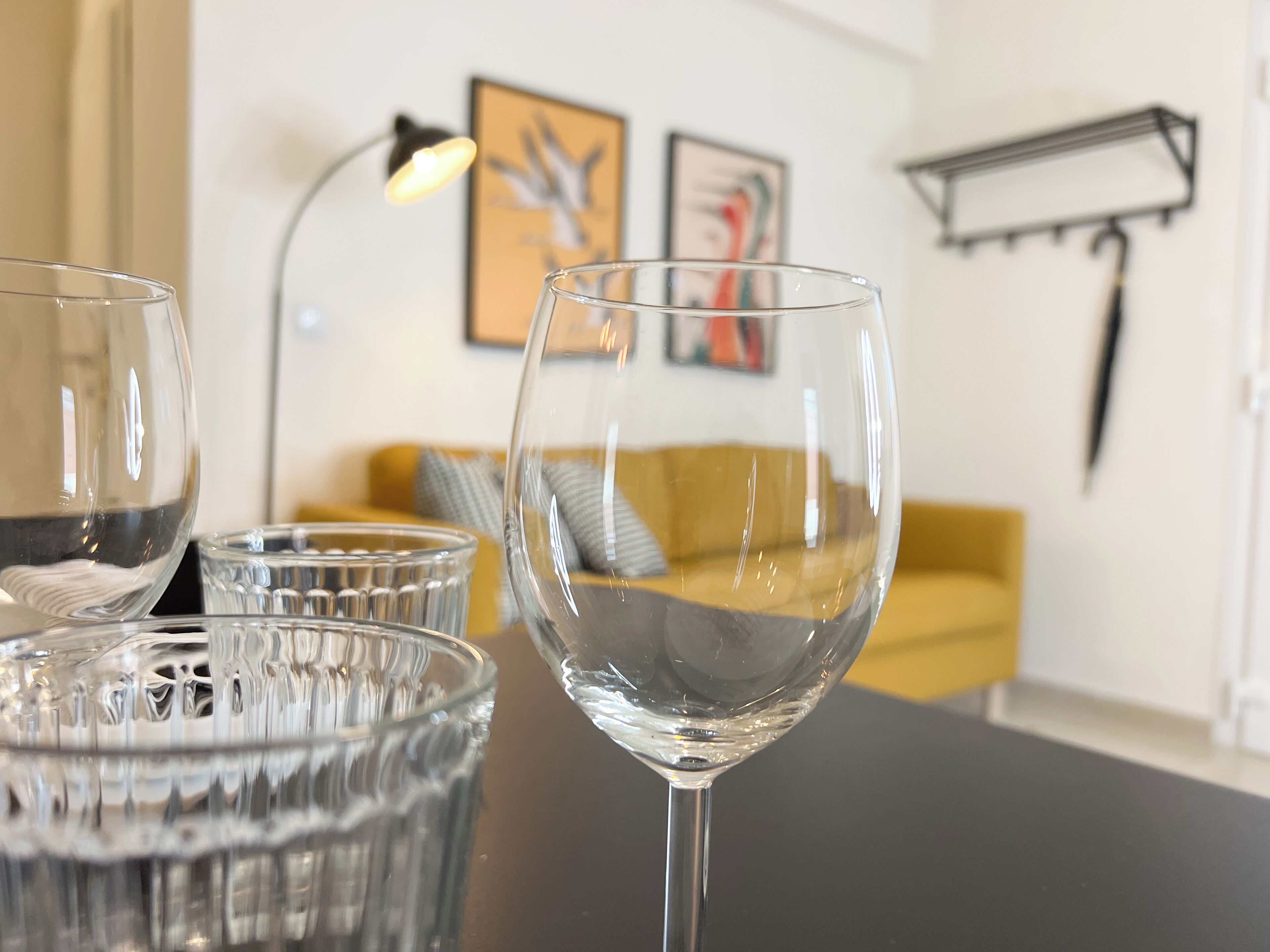 Une table en verre transparent, avec trois verres dessus, une lampe et un canapé jaune en arrière-plan.
