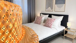 Ein ordentlich bezogenes Bett mit gemusterten Kissen, ein Nachttisch mit Lampe sowie zwei gerahmte Kunstwerke an der Wand.