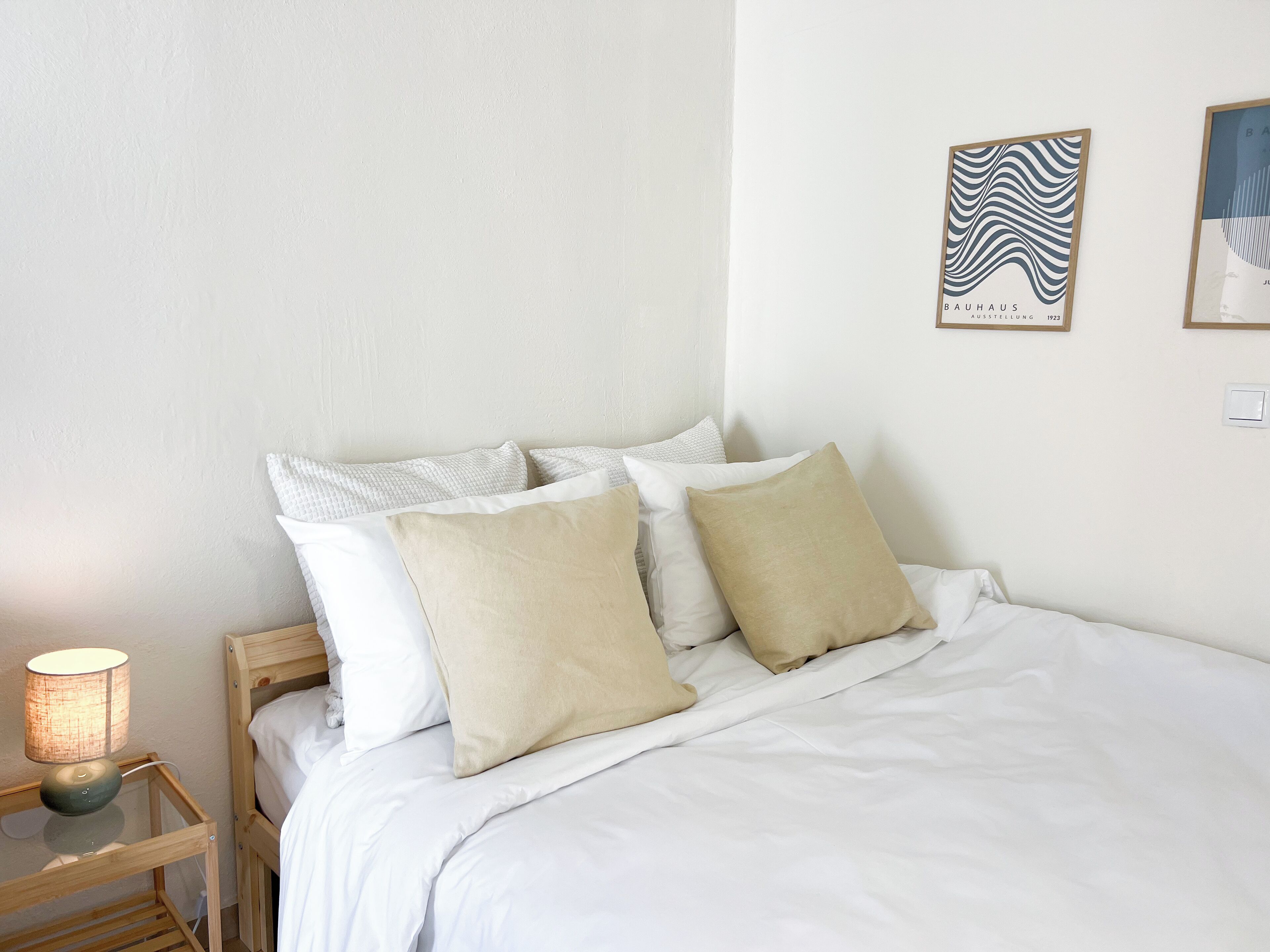 Un lit bien fait, avec des oreillers blancs et beiges, une table de chevet avec une lampe, et deux tableaux encadrés accrochés au mur.