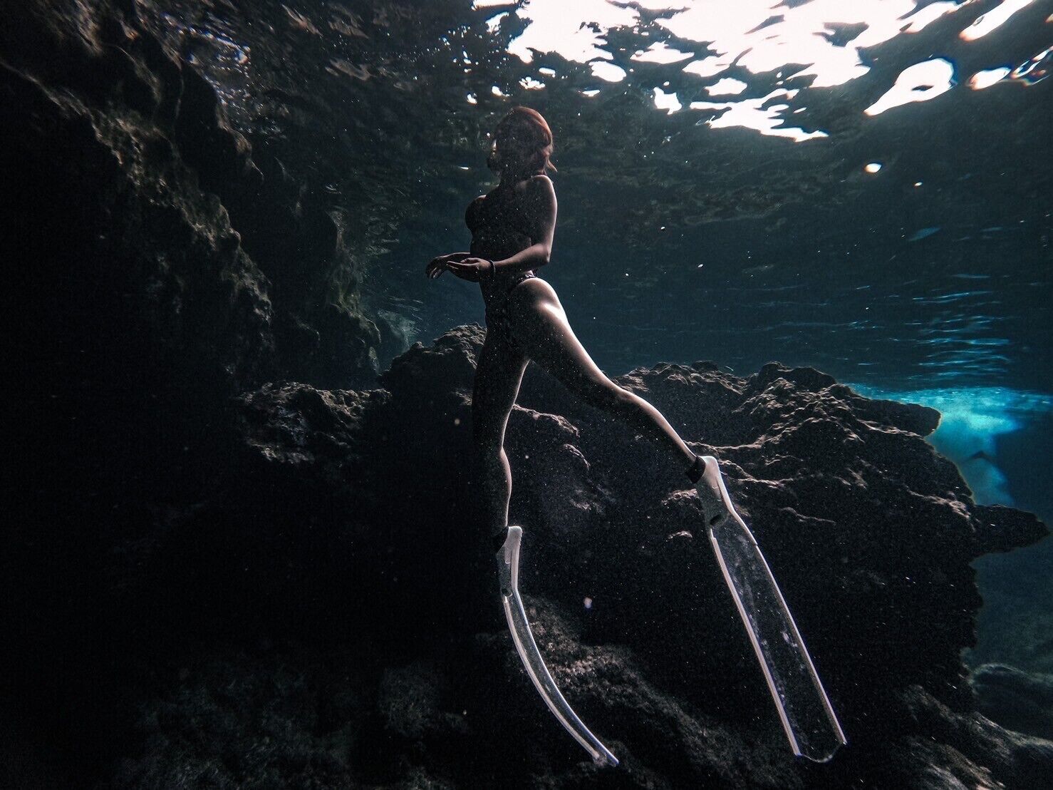 A person in a dark swimsuit standing on a rock with fins, underwater.