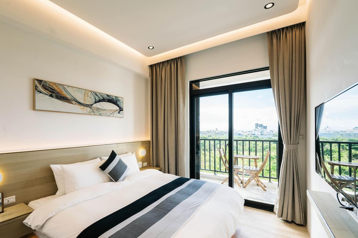 A modern hotel room with a large bed, a city view balcony, and a contemporary art piece on the wall.