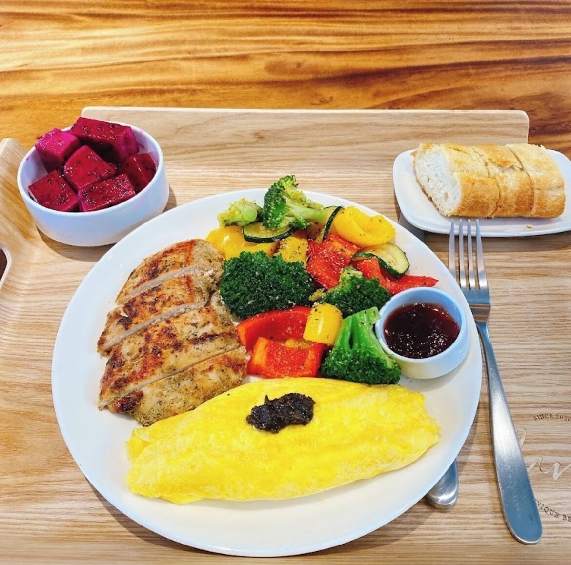 A plate of food including grilled chicken, vegetables, and an omelette, with a side of fruit and bread on a wooden table.