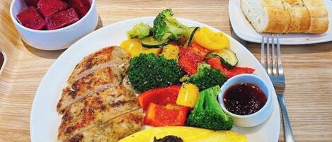 A plate of food including grilled chicken, vegetables, and an omelette, with a side of fruit and bread on a wooden table.