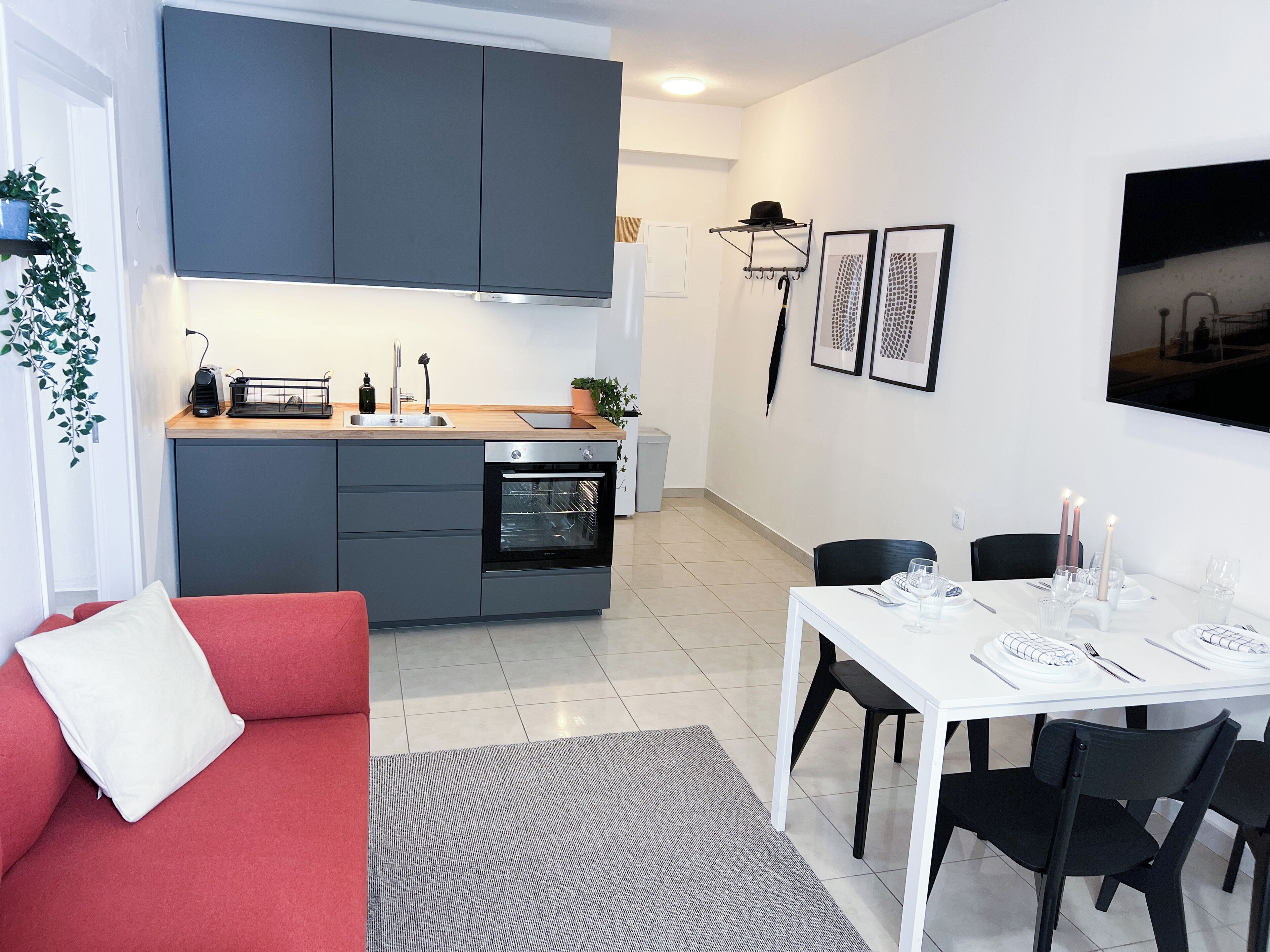A modern kitchen with grey cabinets, a built-in oven, and a sink. There is a dining area with a white table and black chairs.