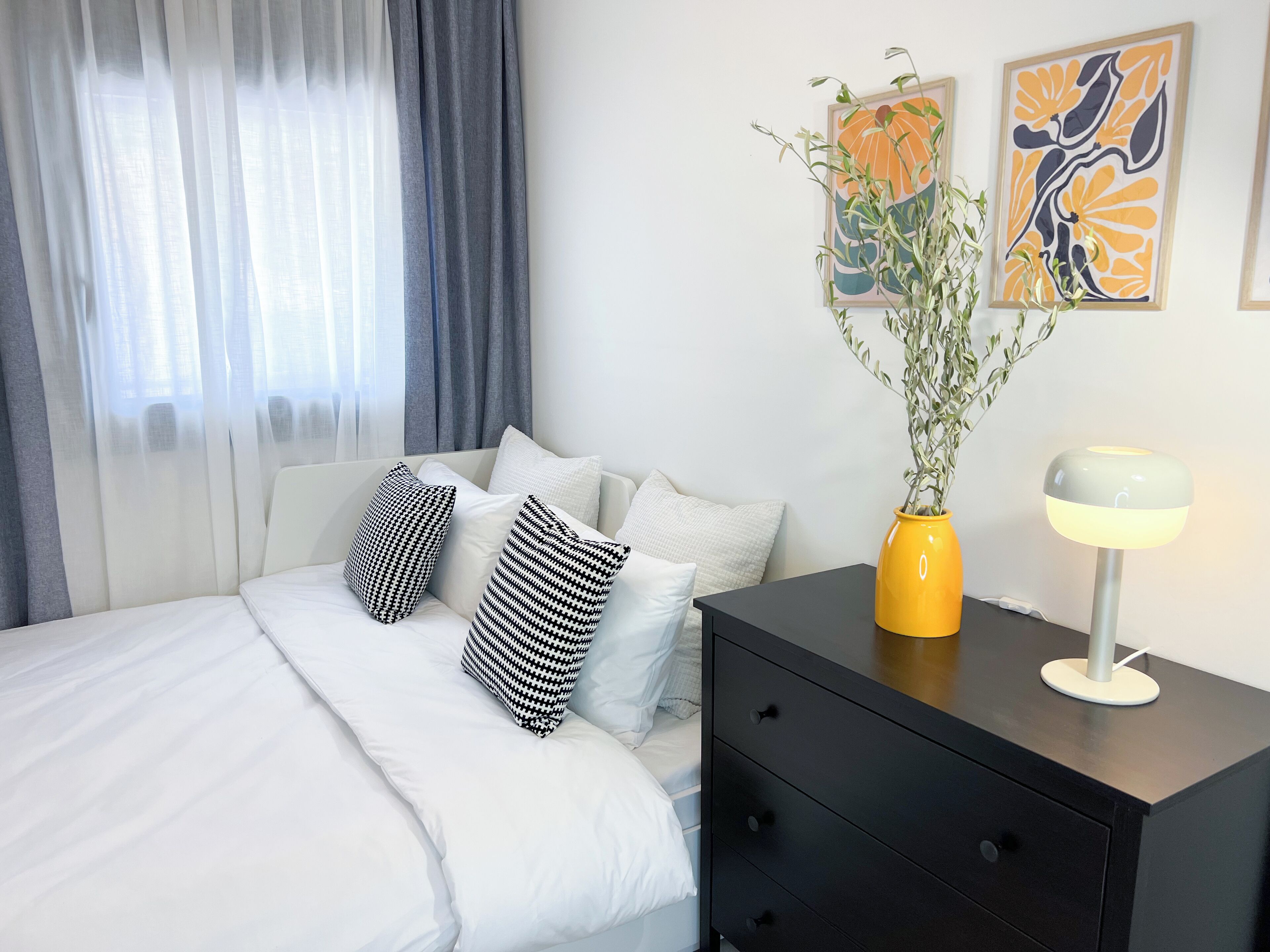 A neatly arranged bedroom with a bed, pillows, a bedside table with a lamp, and two wall art pieces.