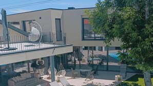 Egy modern, szabadtéri medencetér, napozóágyakkal és egy függőkosárral, melyet egy nagy ablakokkal ellátott épület és egy fa vesz körül.