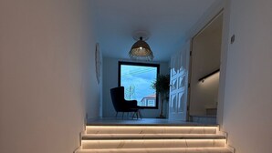 Egy modern lépcső, márvány lépcsőfokkal és beépített világítással, amely egy nagy ablakos, egy székkel és egy függesztett lámpatesttel berendezett helyiségbe vezet.