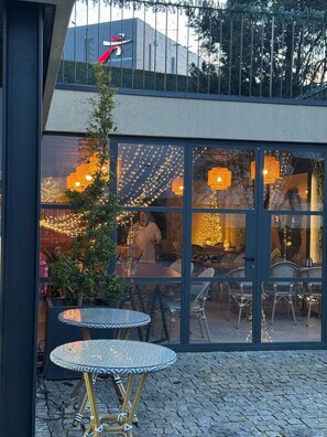 Un bâtiment moderne doté de grandes fenêtres en verre, d’une terrasse avec des tables et des chaises rondes, et d’un éclairage décoratif.