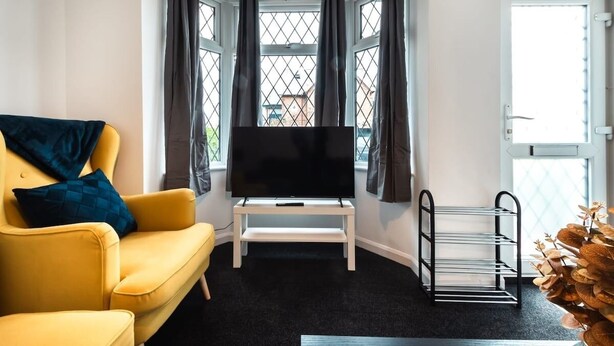 A corner room with a yellow sofa, a flat-screen TV on a white stand, a window with curtains, and a door with a view of the outside. - Central House in Southampton (Freemantle)
