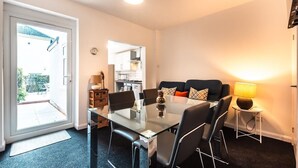 A modern living room with a glass dining table, black chairs, a sofa with cushions, a small side table with a lamp, and a view of the kitchen through an open door. - Central House in Southampton (Freemantle)