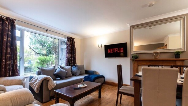 Una sala de estar con sofá, sillones, una mesa de centro, una mesa de comedor con sillas, un televisor montado en la pared y un espejo grande sobre la mesa.