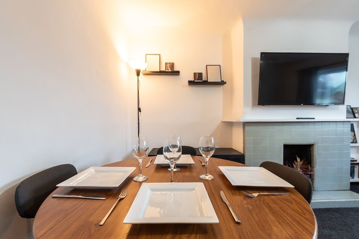 A dining area with a round wooden table set for four, equipped with white plates, cutlery, and wine glasses. There is a flat-screen TV mounted on the wall above the fireplace, and a floor lamp is visible to the left.