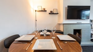 A dining area with a round wooden table set for four, equipped with white plates, cutlery, and wine glasses. There is a flat-screen TV mounted on the wall above the fireplace, and a floor lamp is visible to the left. - Central Flat, Off-Road Parking (Shirley)