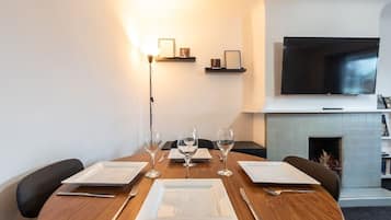 A dining area with a round wooden table set for four, equipped with white plates, cutlery, and wine glasses. There is a flat-screen TV mounted on the wall above the fireplace, and a floor lamp is visible to the left.