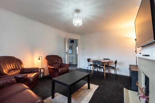 A living room with leather sofas, a coffee table, a chandelier, a dining area with chairs and a table, and a fireplace.