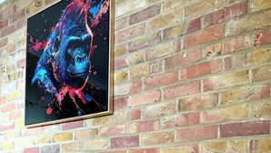 Un mur de briques orné d’un tableau abstrait encadré, une table en verre avec un cadre doré, sur laquelle repose un objet décoratif, et un verre à vin en verre.