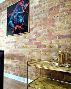 Pared de ladrillo con un cuadro abstracto enmarco, una mesa de vidrio con un marco dorado y un objeto decorativo, y una copa de vino de vidrio.