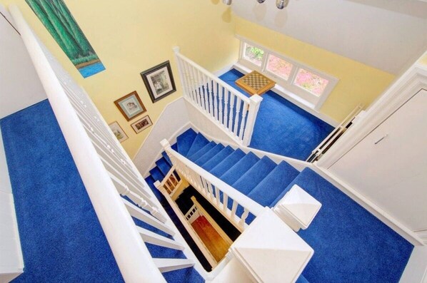 A staircase with blue carpet and white railings, leading to a landing with a window and framed pictures. - Charming Edwardian 5 Bed House | London (Elstree)