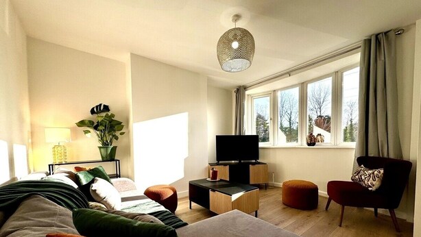 Una sala de estar moderna con sofá, reposapiés y sillón, televisor de pantalla plana, mesita de noche con lámpara y un amplio ventanal con cortinas.