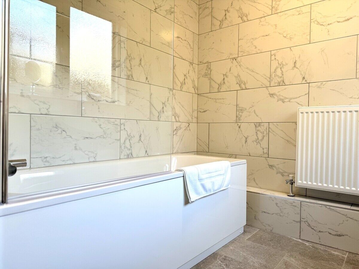 A modern bathroom with a marble-tiled wall, a white bathtub, and a white radiator.