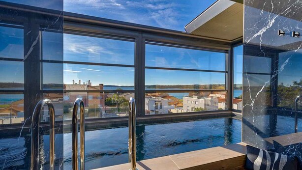 Uma área moderna de piscina interna com paredes de vidro transparente, vista para o horizonte urbano e parede de mármore.