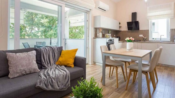 Una sala de estar moderna con sofá, mesa de centro y sillones. Hay un comedor con mesa y sillas. La habitación tiene ventanas amplias y un equipo de aire acondicionado en la pared.
