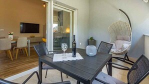 Un comedor con una mesa dispuesta para dos personas, una botella de vino y una copa. Hay una silla de mimbre y una silla acolchada con asiento colgante. En el fondo, un televisor está montado en la pared.