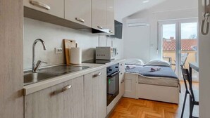 A compact kitchen with a sink, microwave, and oven, a small dining area with a table and chairs, and a bed with a view of the outside.