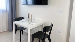 Un coin repas avec une table blanche, deux chaises noires, un téléviseur à écran plat fixé au mur et un chandelier avec des bougies.