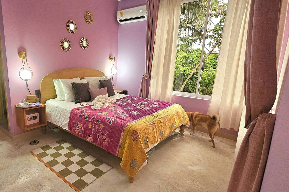 A bedroom with a bed, bedside tables, a lamp, a window with curtains, and a small dog.