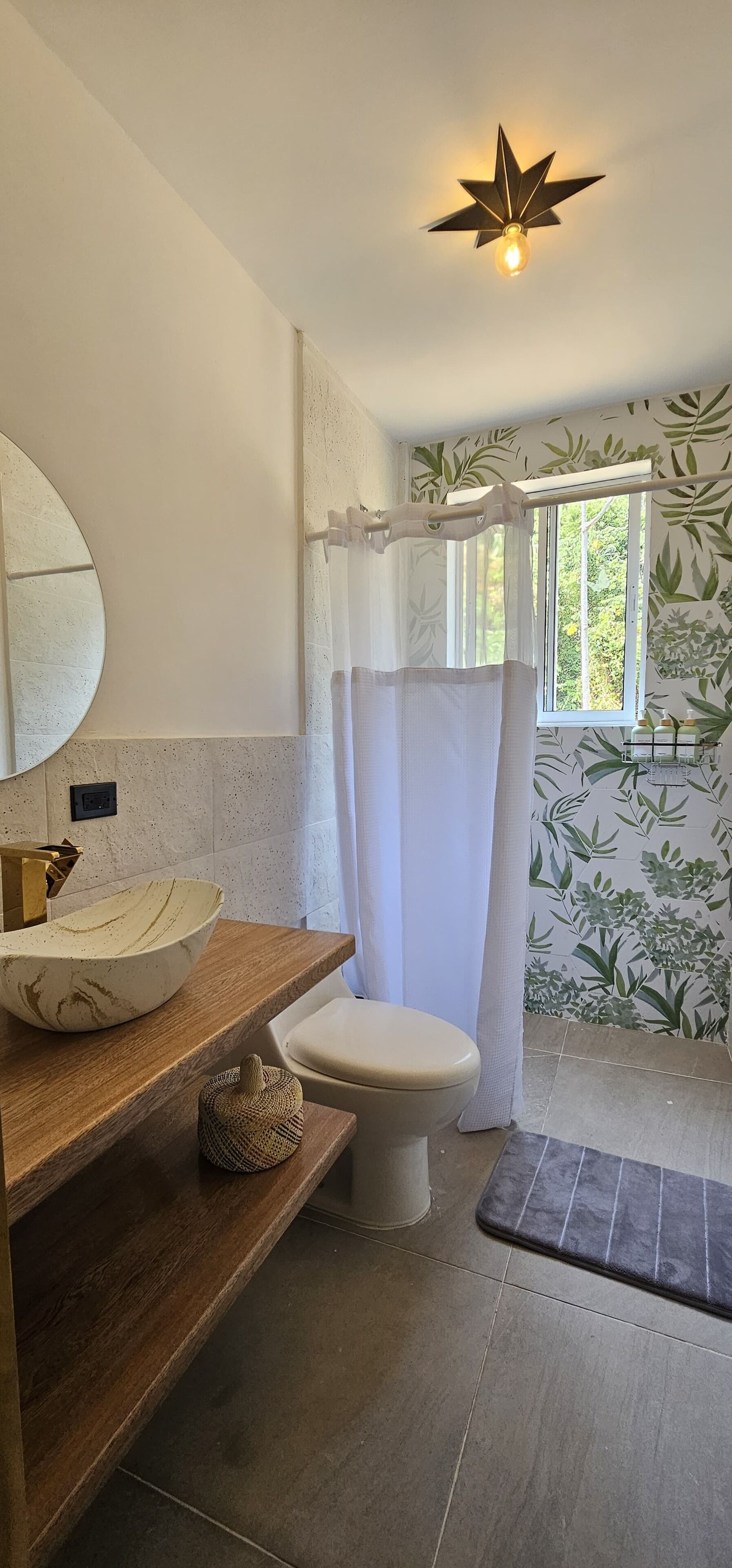 A bathroom with a wooden vanity, a round mirror, a white toilet, a shower curtain, and a window with a view of greenery.