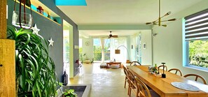 Une salle à manger spacieuse avec une longue table et des chaises en bois, un ventilateur de plafond et une vue sur l’extérieur grâce à de grandes fenêtres.