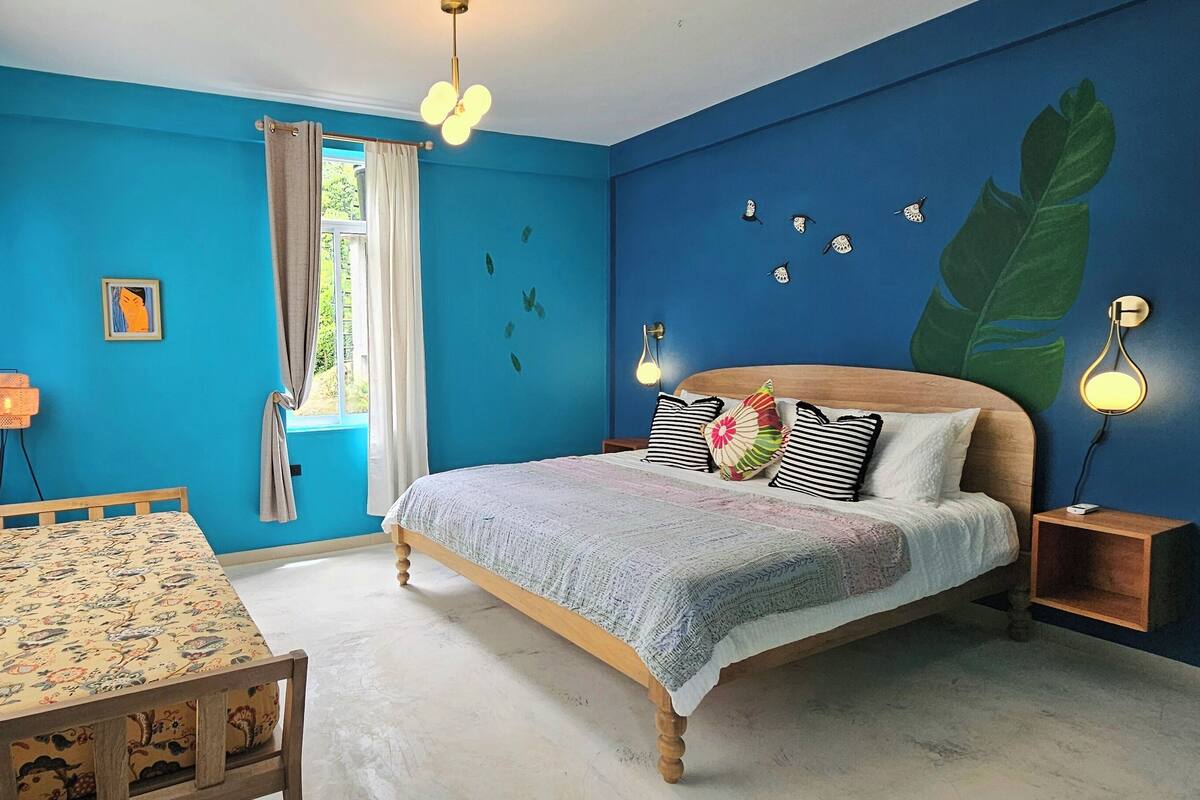 A bedroom with a bed, a table, and a chair. There is a window with curtains, a wall with a leaf mural, and a wall-mounted light fixture.