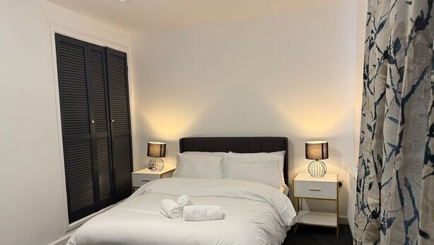 A neatly made bed with white linens and two rolled towels, flanked by two bedside tables with lamps, in a room with a dark headboard and a window with closed blinds. - Hideaway 4-Bed House in Bridgend" (Bridgend County Borough)
