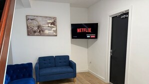 A living room with a blue sofa, a television mounted on the wall displaying 'NETFLIX', and a door to the right. - Hideaway 4-Bed House in Bridgend" (Bridgend County Borough)