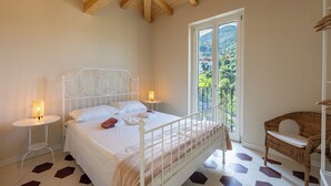 Une chambre à coucher avec un lit, des tables de chevet, un fauteuil en osier et un balcon donnant sur un paysage verdoyant.