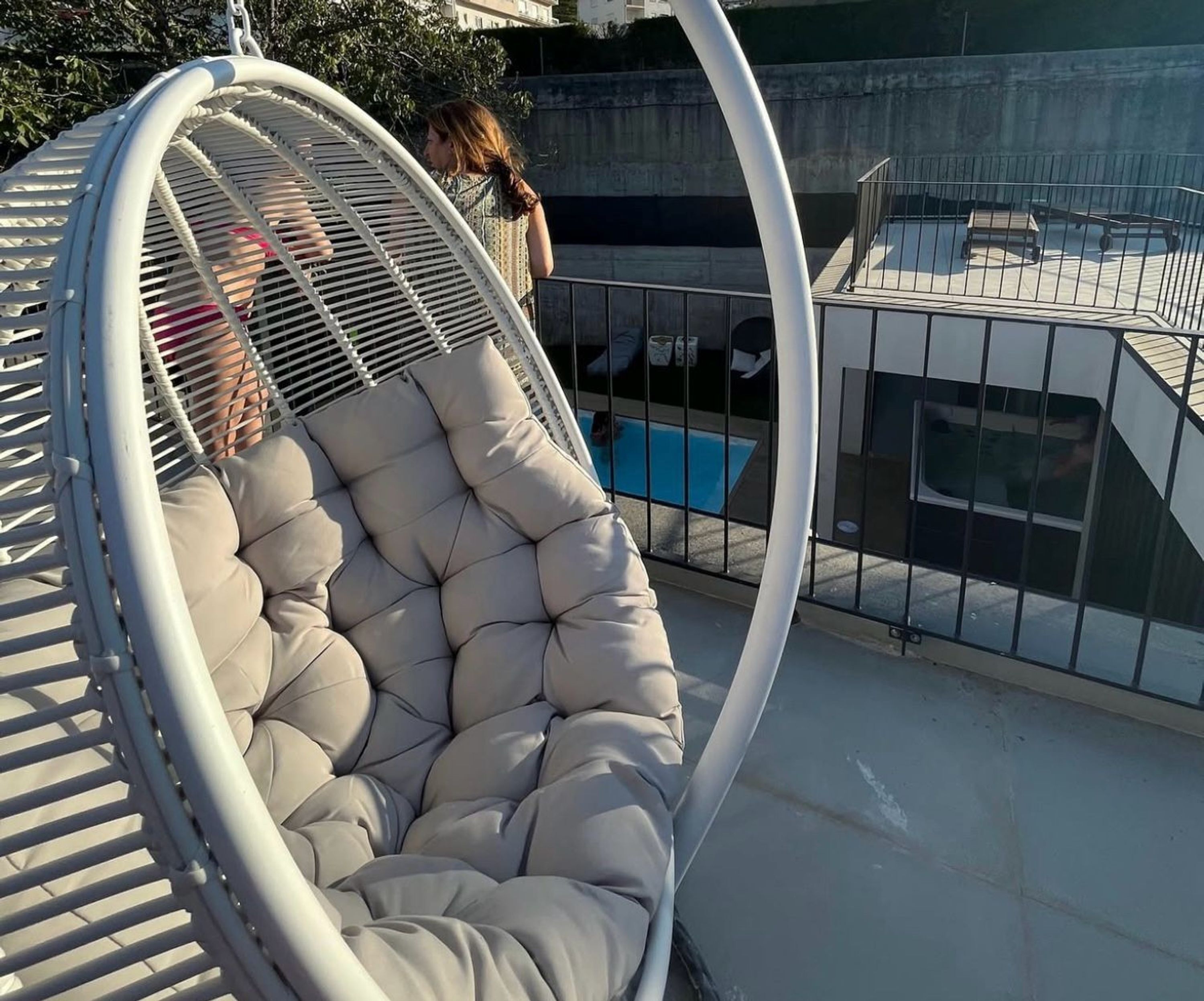 A hanging chair with a cushioned seat and a metal frame, overlooking a swimming pool area with a concrete wall and a metal fence.