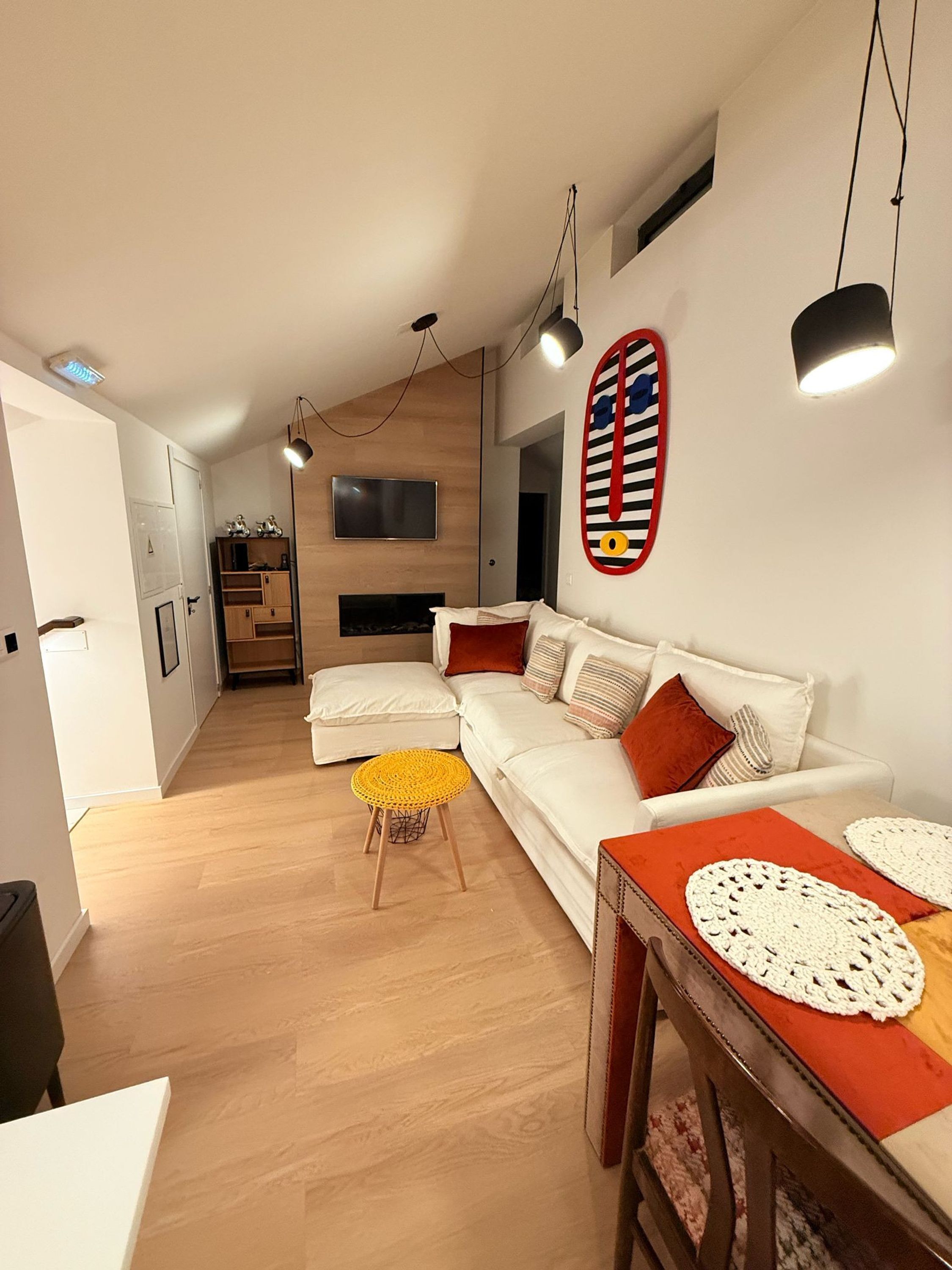 Una sala moderna con un sofá blanco, una mesa de centro de madera y un taburete amarillo. Hay un televisor montado en la pared y una chimenea al fondo.