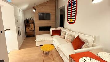 Una sala moderna con un sofá blanco, una mesa de centro de madera y un taburete amarillo. Hay un televisor montado en la pared y una chimenea al fondo.