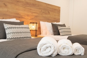 A neatly made bed with grey bedding, two patterned pillows, a wooden headboard, a bedside lamp, and rolled white towels.