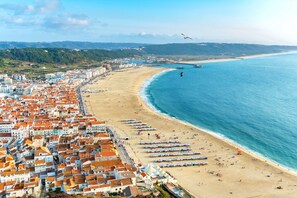 A coastal cityscape with a beach lined with rows of beach umbrellas and tents, a clear blue sea, and a dense urban area with terracotta-roofed buildings.