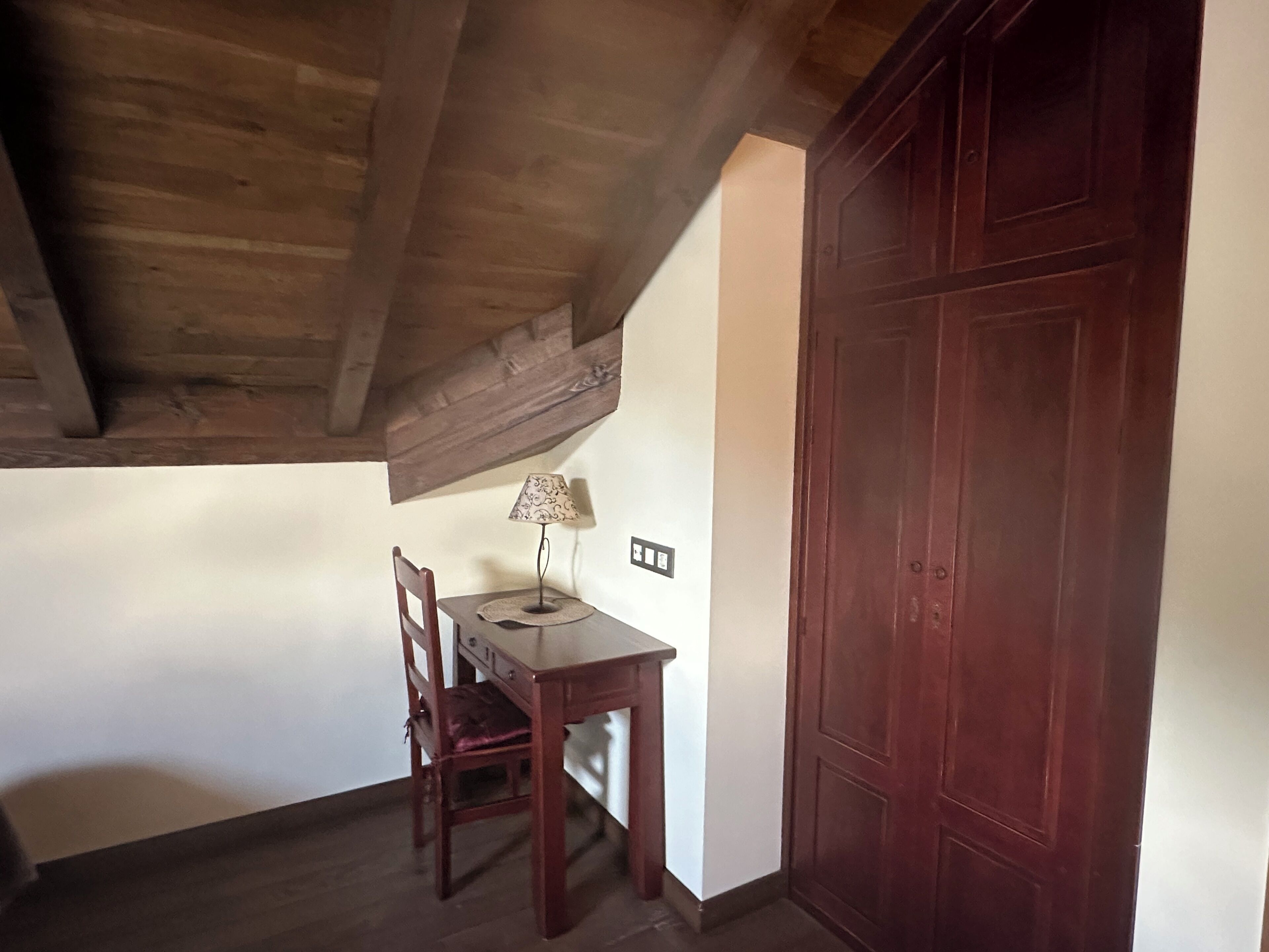 A room with a wooden ceiling, a desk with a lamp, a chair, and a large wooden door.