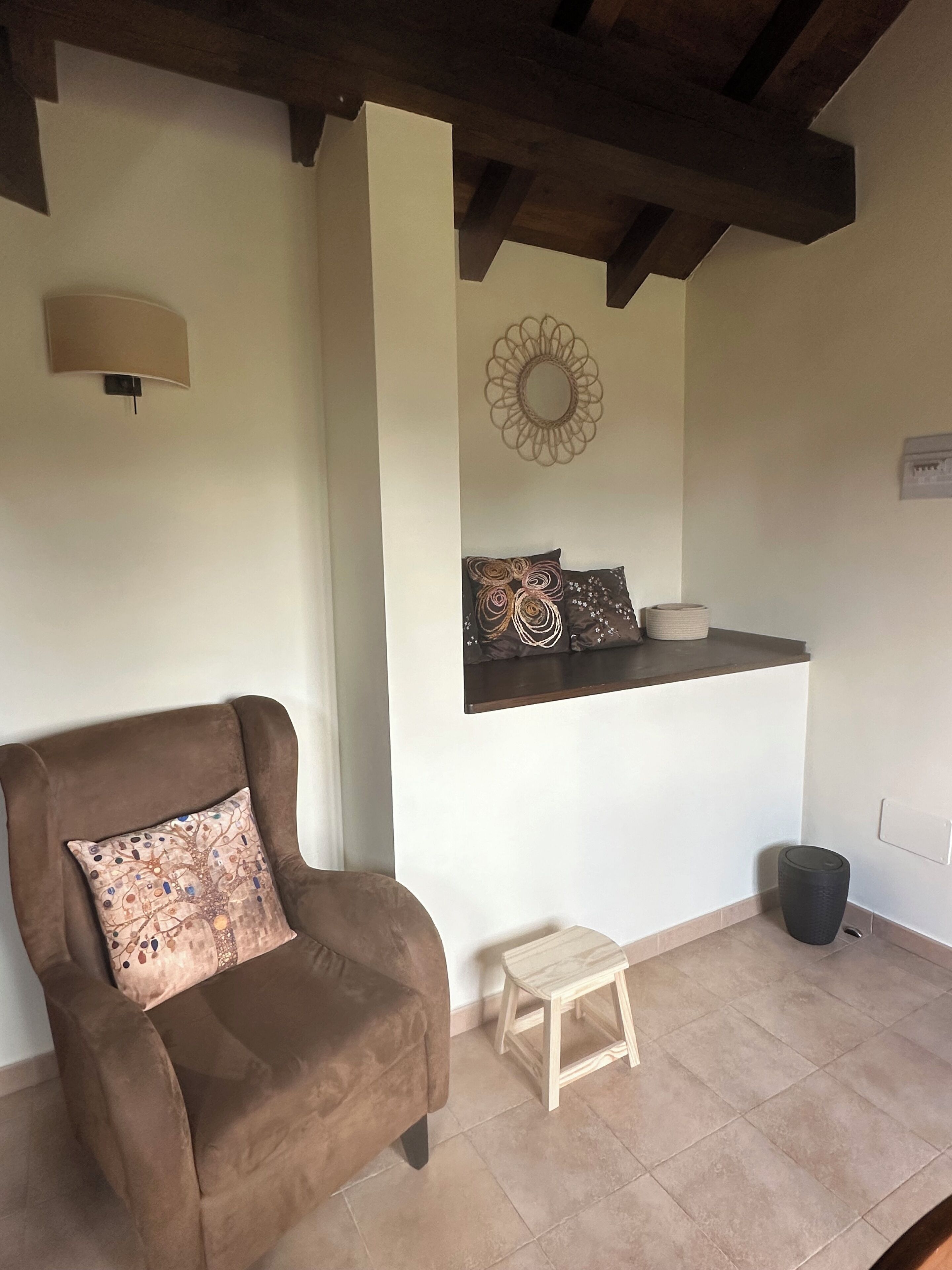 Una habitación acogedora con un sillón marrón, un taburete de madera, un adorno en la pared y una estantería con un espejo y almohadones.