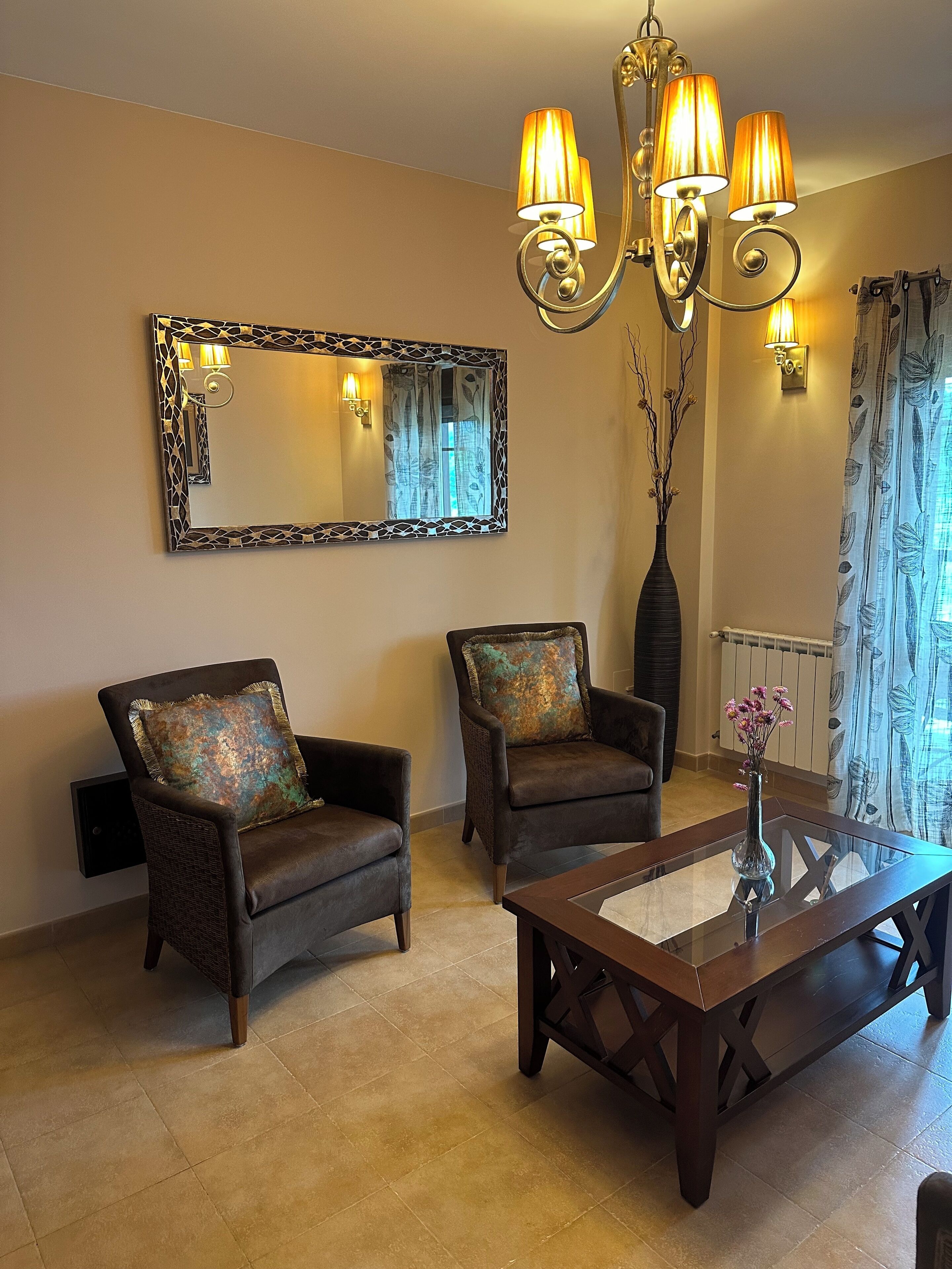 A living room with two armchairs, a glass coffee table, a chandelier, a large mirror, and a vase with flowers.