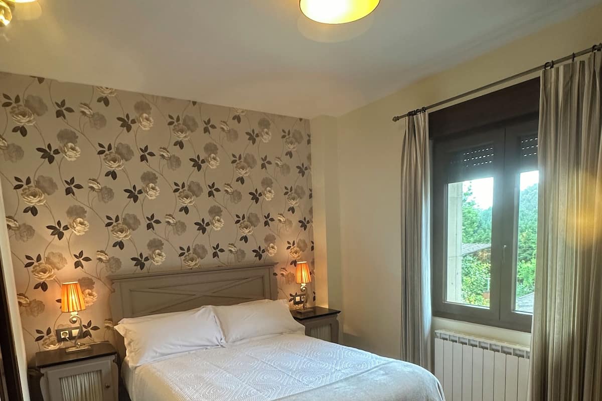 A bedroom with a bed, two bedside lamps, a window with curtains, and floral wallpaper.