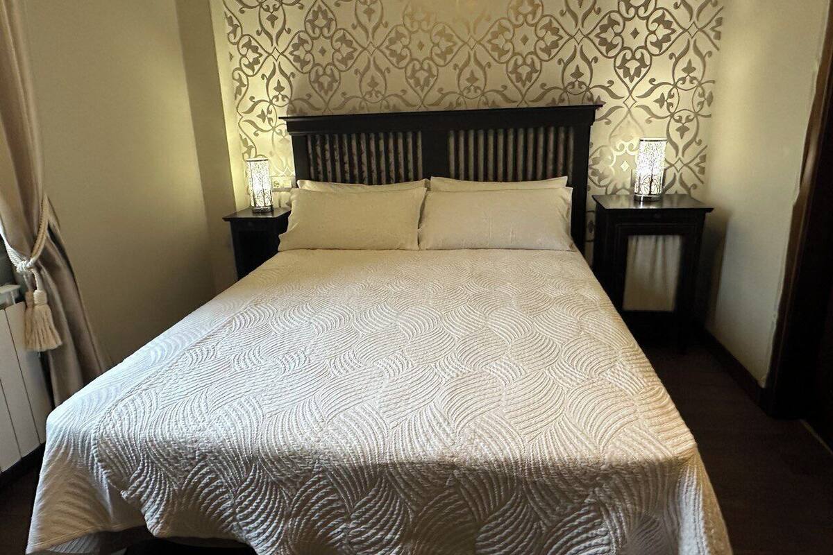 A neatly made bed with a patterned bedspread, two bedside tables with lamps, and a patterned wallpaper on the wall.