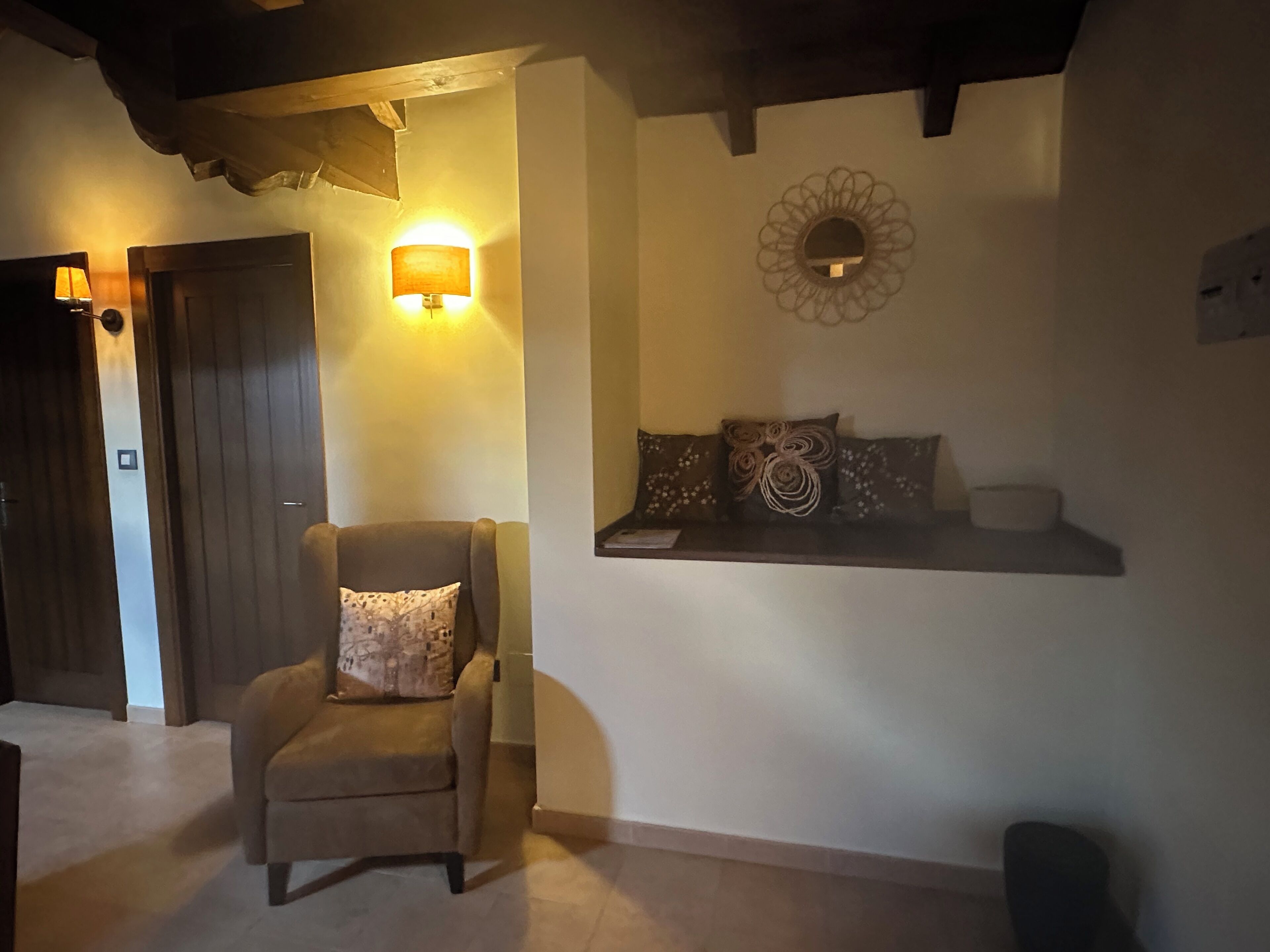 A cozy room with a wooden ceiling, a cushioned armchair, a wall-mounted shelf with decorative pillows, and a wall-mounted light fixture.