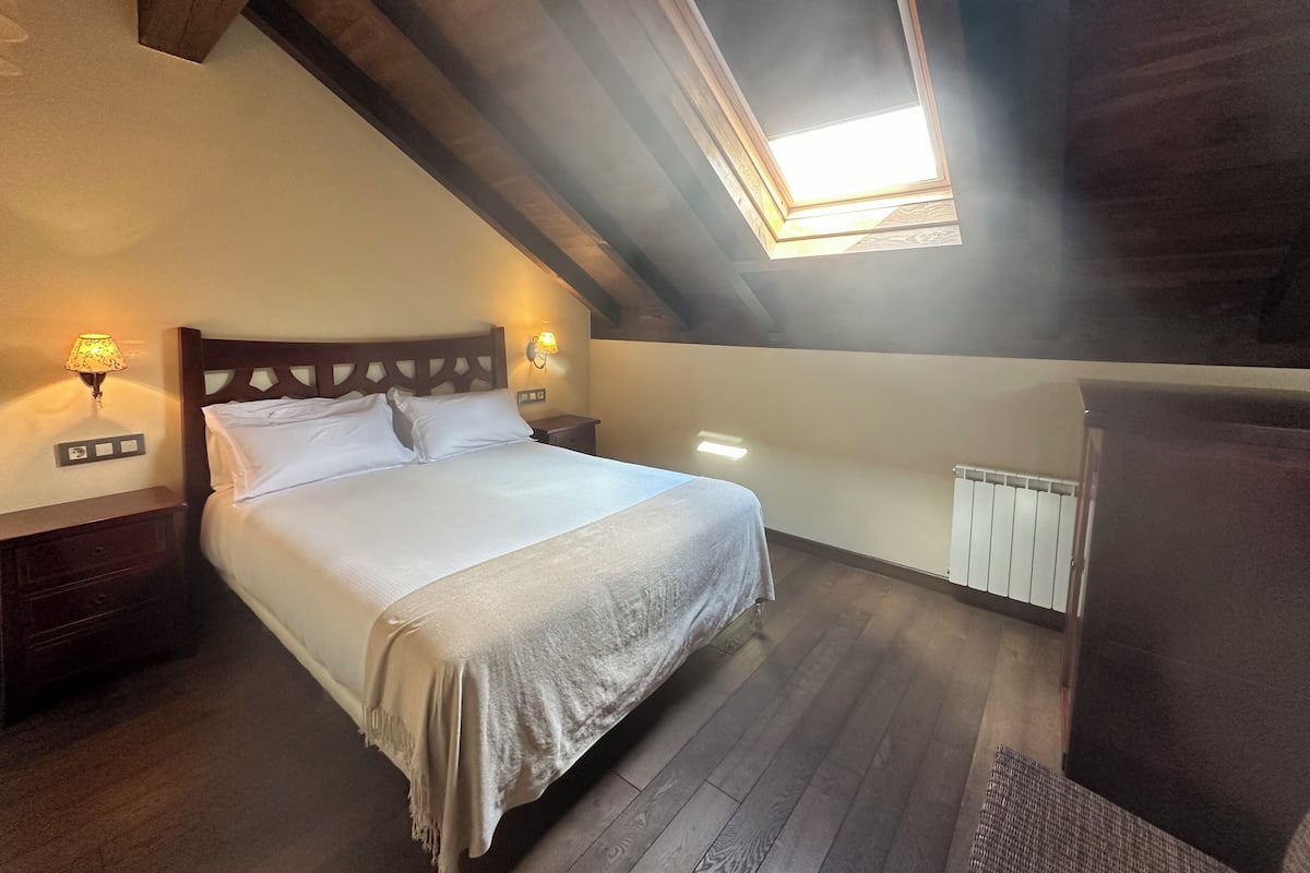 A bedroom with a wooden ceiling, a skylight, a bed with white linens, two bedside lamps, and a radiator.